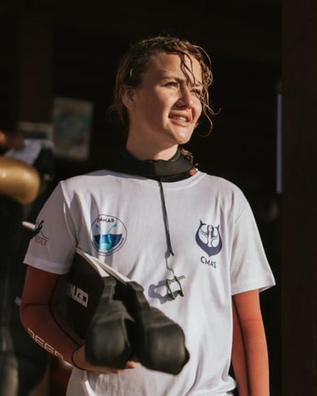 A young white woman wearing a wetsuit and carrying fins walks through a doorway