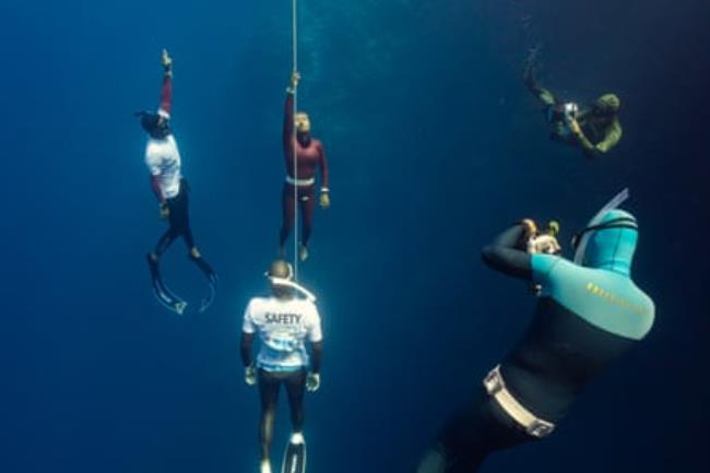 A freediver in a burgundy wetsuit swims to the surface while holding on to a rope as two other divers accompany her and two photographers take pics   