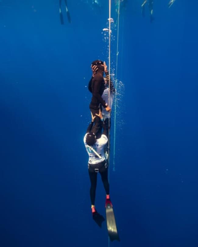 An unconscious diver in a black wetsuit is supported vertically in the water by two safety divers in white shirts next to a safety rope